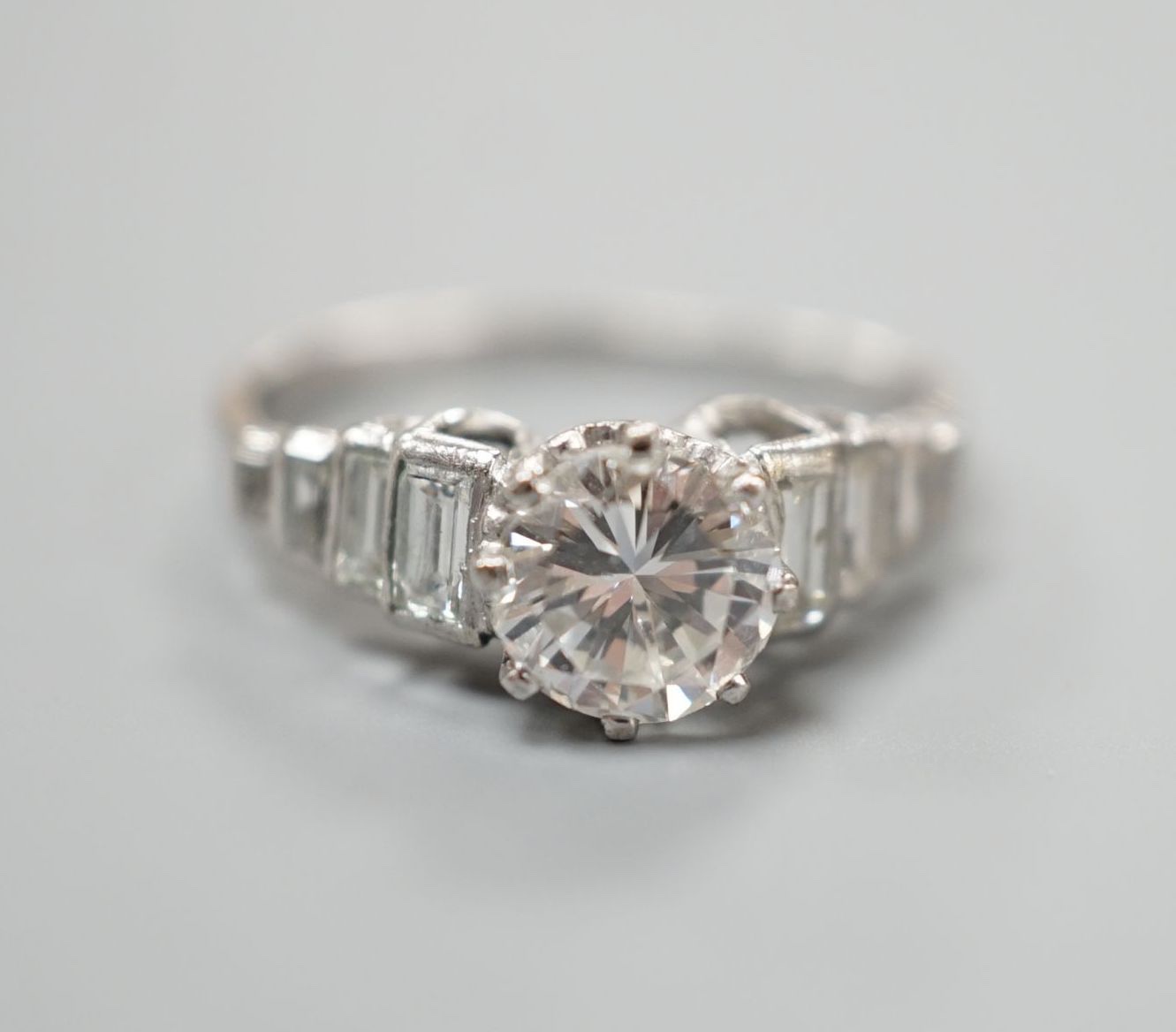 A white metal (stamped Plat) and single stone diamond ring, with graduated eight stone baguette cut diamond set shoulders, size K/L, gross weight 3.5 grams